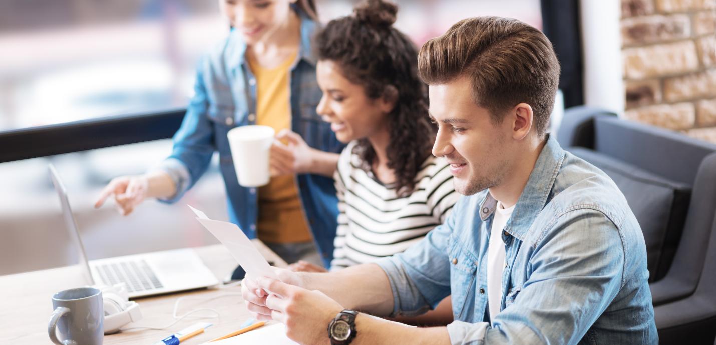 A young man and two young ladies sitting at a desk with a laptop and some paperwork